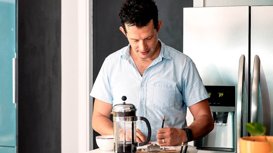 Jon Hagar writes notes on the counter in his kitchen, with a coffee press and cup near him and the refrigerator behind him.