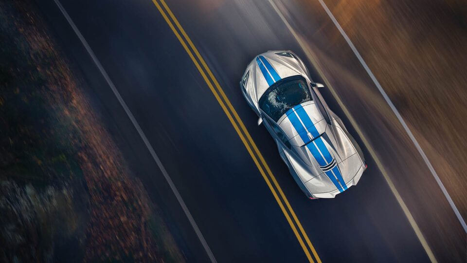 A gray Corvette E-Ray with blue racing stripes seen from overhead on a two-lane road.