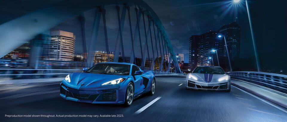 A blue Corvette E-Ray drives over a bridge at night next to a gray E-Ray with blue racing stripes.