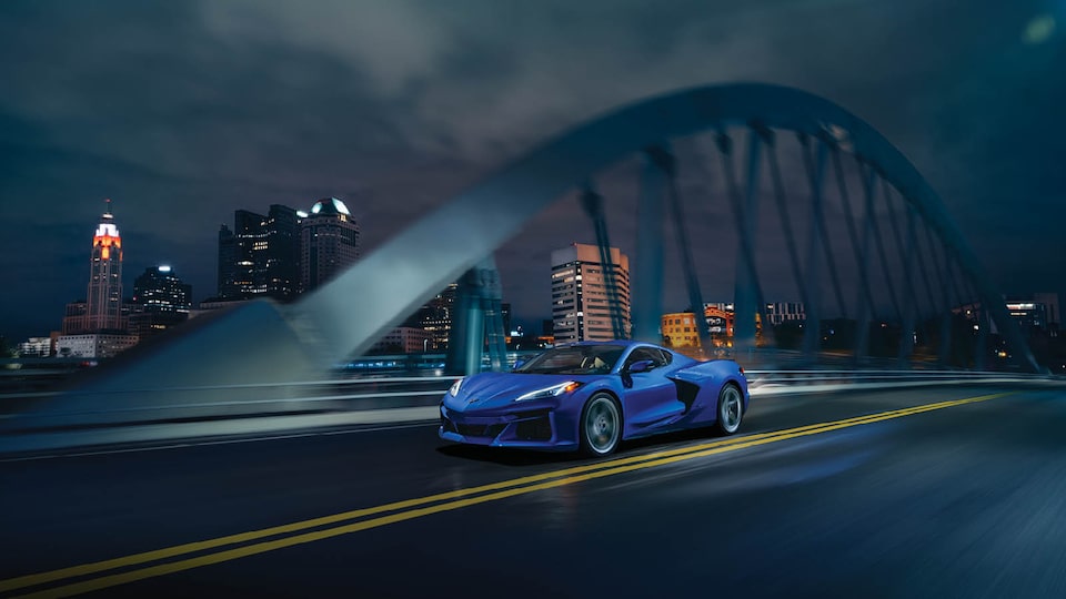 A blue Corvette E-Ray drives over a bridge at night, with tall buildings lit up in the background.