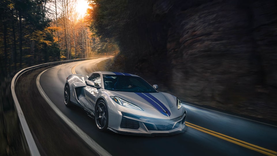 A gray Corvette E-Ray with blue racing stripes drives on a winding, tree-lined road.