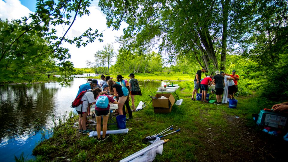 Students from the University of Maine Stormwater Management and Research Team stand in groups with their equipment next to a pond.