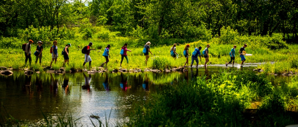 A group of students from the University of Maine Stormwater Management and Research Team walk in a single-file line next to a pond.