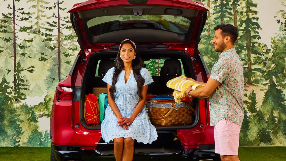 En un set decorado creado para lucir como un área de picnic en el bosque, una mujer está sentada en el maletero de una Chevy Blazer color Rojo Cereza Tintcoat al lado de una canasta de mimbre mientras un hombre está parado al lado del auto con una manta doblada para picnic.