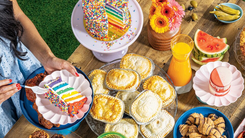 Comida de diferentes colores sobre una mesa de madera para picnic. Una mujer parada al lado de la mesa con un plato y una porción de torta con colores del arco iris y un tenedor.