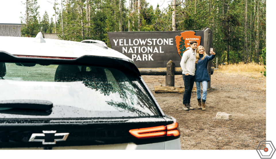 Dos personas se toman una selfie juntas, paradas delante del cartel de entrada del Parque Nacional Yellowstone, con una Bolt EUV blanca de fondo.