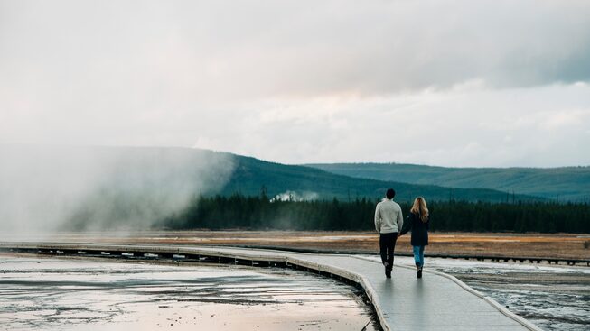 Two people walk on a boardwalk in Yellowstone with steam rising from a hot pool to their left.