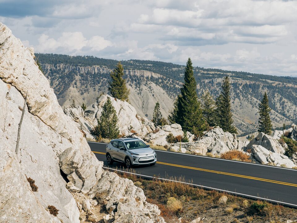 Una Bolt EUV color blanco rodea una curva en el Parque Nacional Yellowstone y emerge por detrás de una formación rocosa, con un fondo boscoso.