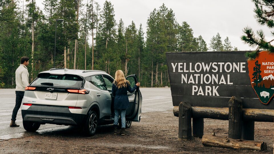 Dos personas subiéndose a una Bolt EUV blanca estacionada al lado de un cartel de entrada de Yellowstone National Park.