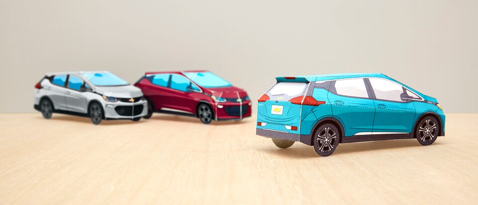 Three completed paper models of the Bolt EV in Silver Ice Metallic, Cajun Red, and Oasis Blue sit on a tabletop.