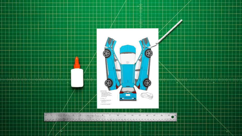 A paper template for the model Bolt EV sits on a green artist mat with an artist knife, a bottle of glue and a ruler. In the video, the paper car is cut out and assembled and drives off the mat.