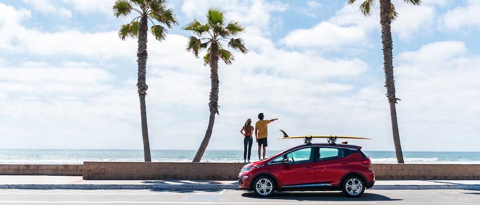 A red Chevrolet Bolt EV with a surfboard strapped to a roof rack sits on a road in front of three palm trees and the beach and ocean beyond the trees. Two people stand on a short wall behind the Bolt EV pointing out at the water.