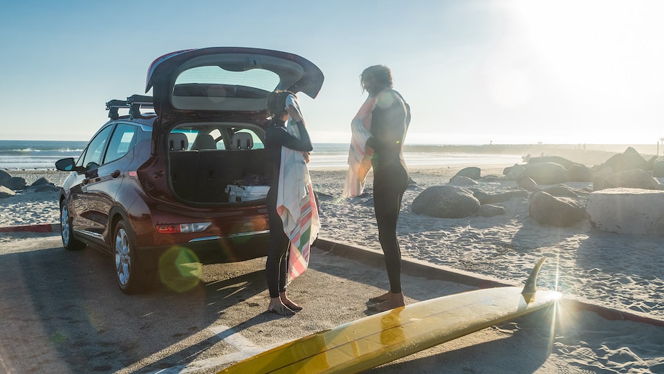 Two people in wetsuits walk from a park- ing lot out onto an ocean beach carrying a  surfboard. A red Chevrolet Bolt EV sits in  the parking lot