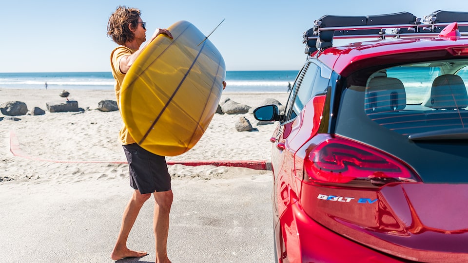 A man holding a surfboard prepares to load it onto the roof rack on a red Chevrolet Bolt EV. The beach is in the background.