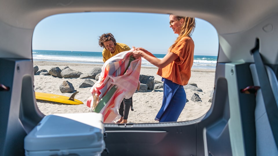 With a viewpoint from inside a Bolt EV looking out the raised hatch in back, a woman and a man unload surfing gear onto a beach. The ocean is in the background.