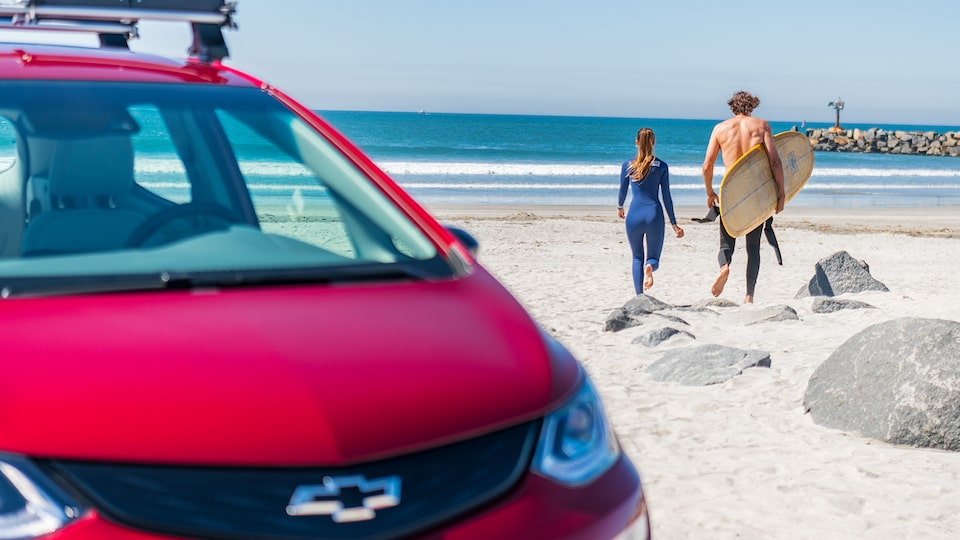 A woman and a man holding a surfboad walk across the beach toward the ocean. A red Chevrolet Bolt EV is in the foreground.