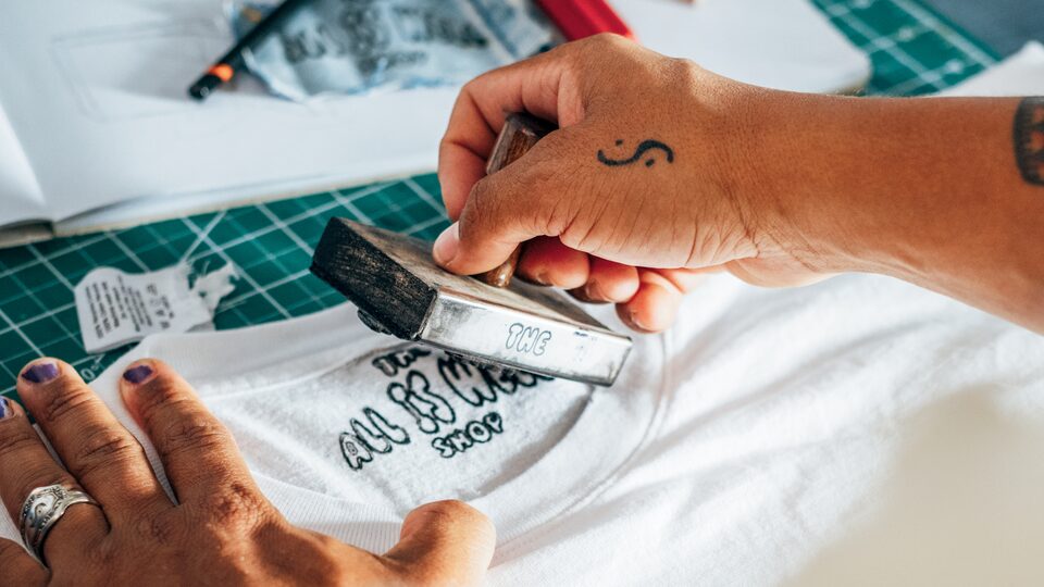 A pair of hands using an ink stamp  to stamp the "All Is Well" logo on the  inside neck of a t-shirt.