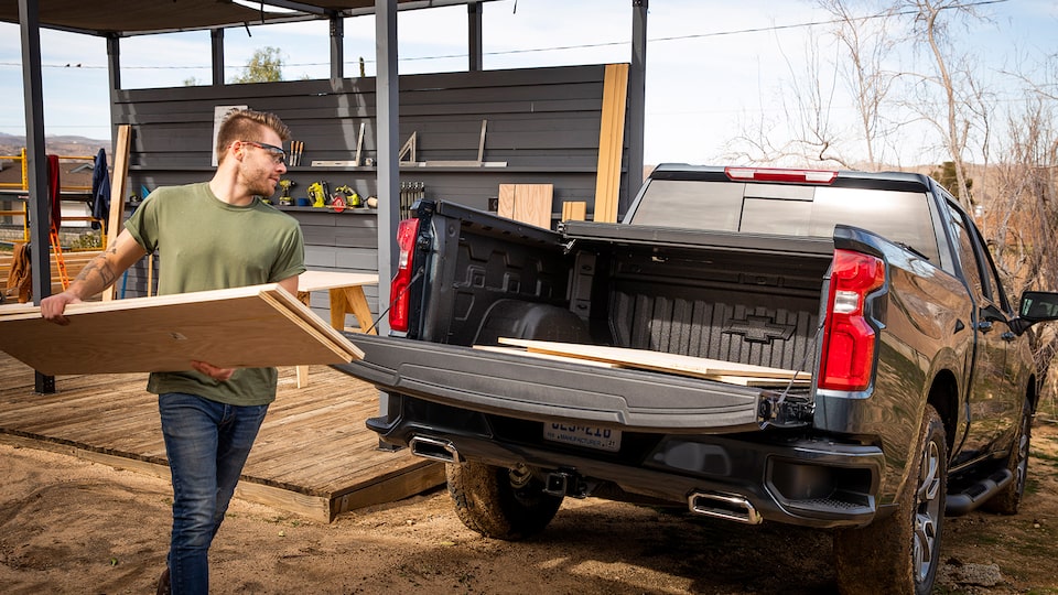 Un hombre con ropa de trabajo y gafas de seguridad carga láminas de madera contrachapada en la caja abierta de una camioneta pickup Chevy Silverado en Negro.