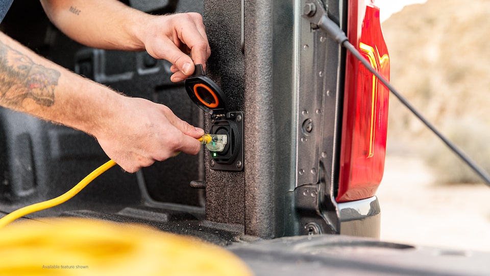 Se ven las manos de un hombre enchufando un cable de extensión amarillo en el enchufe de la parte trasera de la caja de una camioneta pickup Chevy Silverado en Negro.