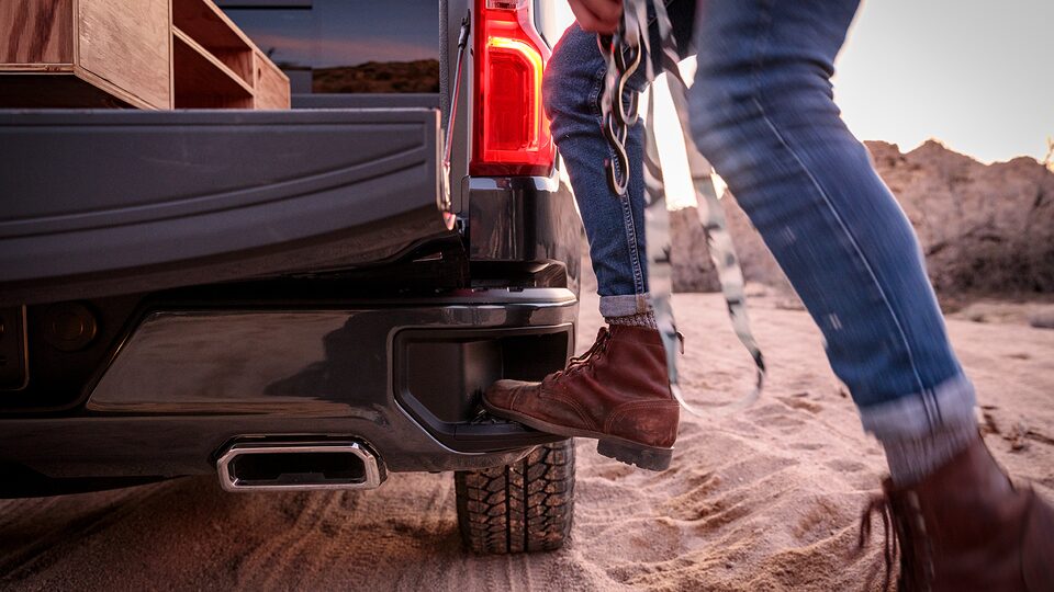 Se ve la parte inferior de las piernas de un hombre con botas mientras sube al escalón de la defensa negra de una camioneta pickup Chevy Silverado en Negro.