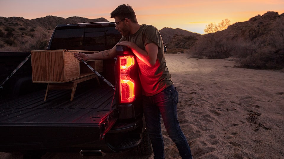 Un hombre con ropa de trabajo se arrodilla en la caja abierta de una camioneta pickup Chevy Silverado en Negro. Está ajustando algunas maderas en la caja.