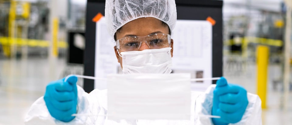A person wearing a face mask, hair net, and gloves displays a face mask by holding it by the elastic on both sides.