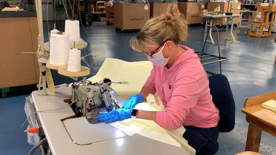 A woman wearing a face mask and gloves sits at a workstation in a plant using an industrial sewing machine with spools of thread behind her.