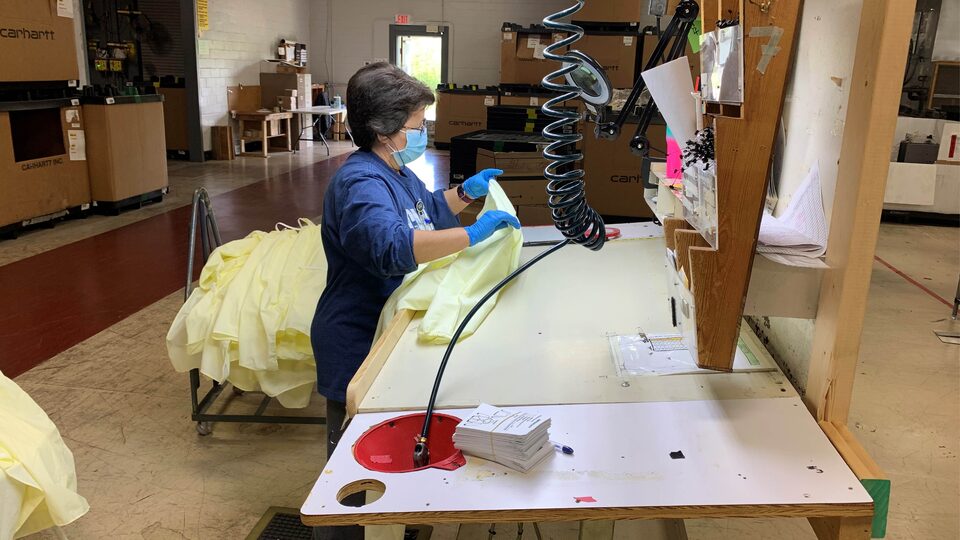 A woman wearing a face mask and gloves works at a large industrial workstation. She’s holding a protective gown and there’s a stack of gowns behind her.
