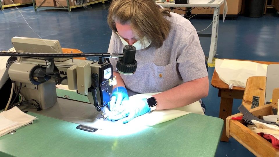 A woman wearing a face mask and gloves uses a bright lamp while she works at an industrial sewing machine in a manufacturing plant.