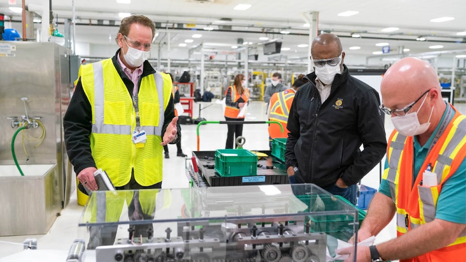 Three men wearing face masks and bright construction vests stand around a piece of equipment in a manufacturing plant. Other people in construction vests are in the background.