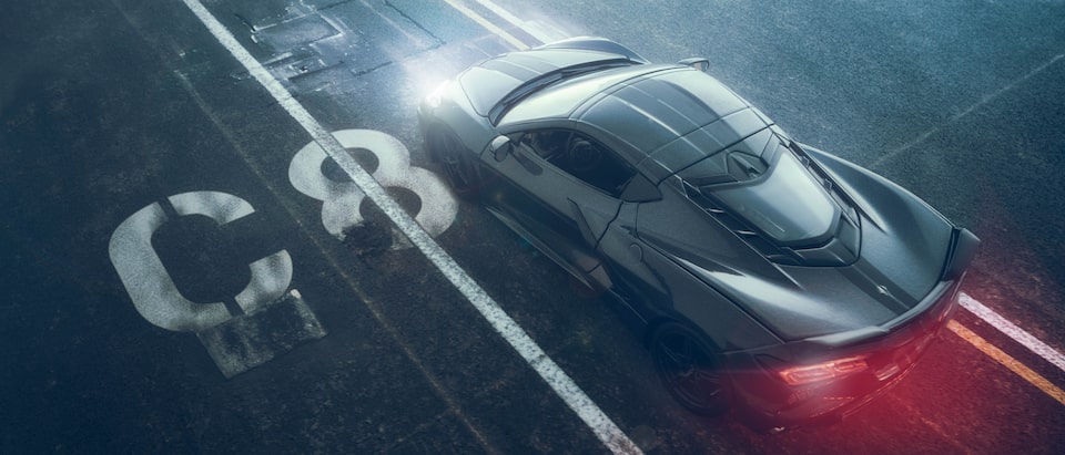A miniature black Corvette Stingray seen from above, with a red glow coming from its taillights, appears to be moving on a backdrop of black pavement with lane markings and a white “C8” stencil next to the car.