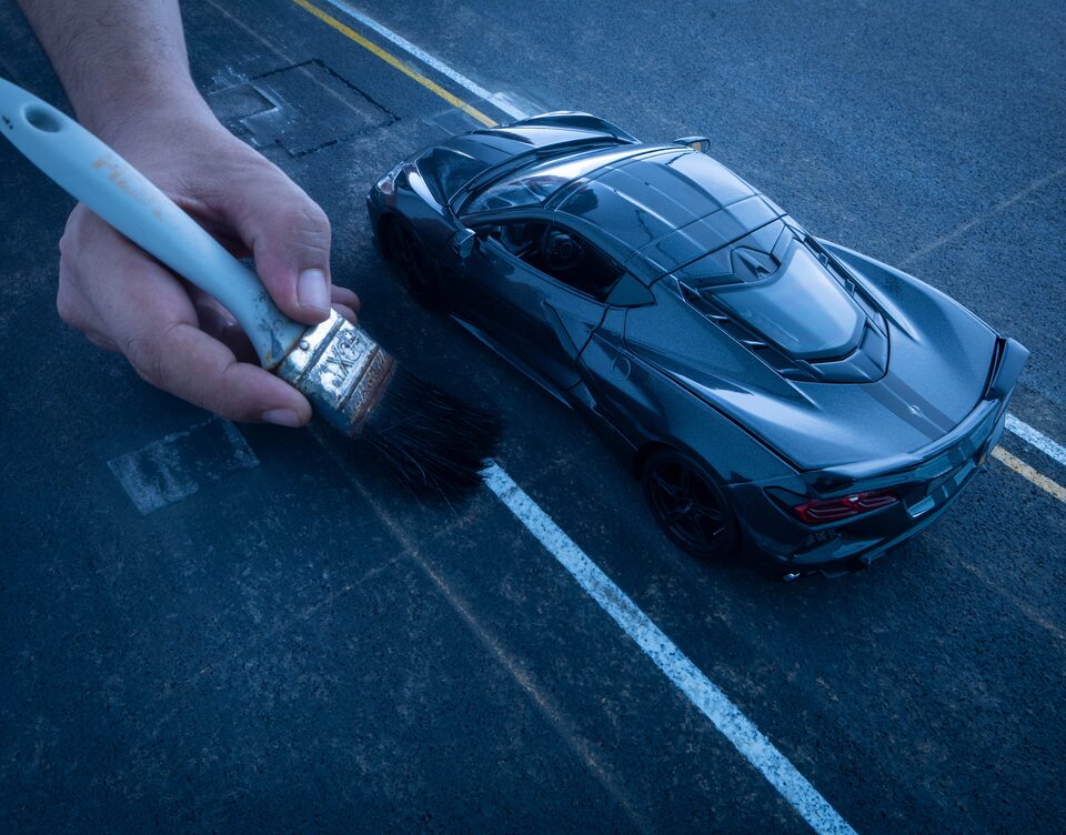 Una mano que sostiene un pincel pinta una línea blanca sobre una calle de fondo, debajo de un Corvette negro en miniatura.