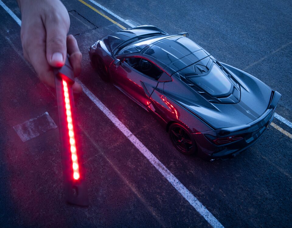 Una mano sostiene una luz roja que ilumina el lado de un Corvette negro en miniatura.