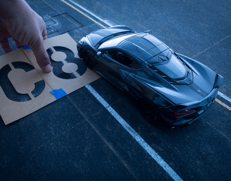 Una mano que sostiene un esténcil de "C8" coloca este en la calle de fondo, debajo de un Corvette negro en miniatura.