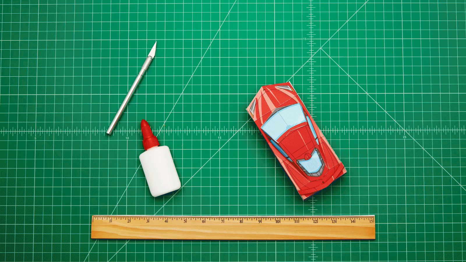 Un gif animado de un modelo rojo de un Corvette hecho de papel moviéndose por el tablero verde de un artista junto a un cuchillo para artes, una pequeña botella de pegamento y una regla.