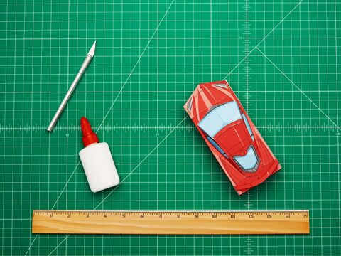 Un gif animado de un modelo rojo de un Corvette hecho de papel moviéndose por el tablero verde de un artista junto a un cuchillo para artes, una pequeña botella de pegamento y una regla.