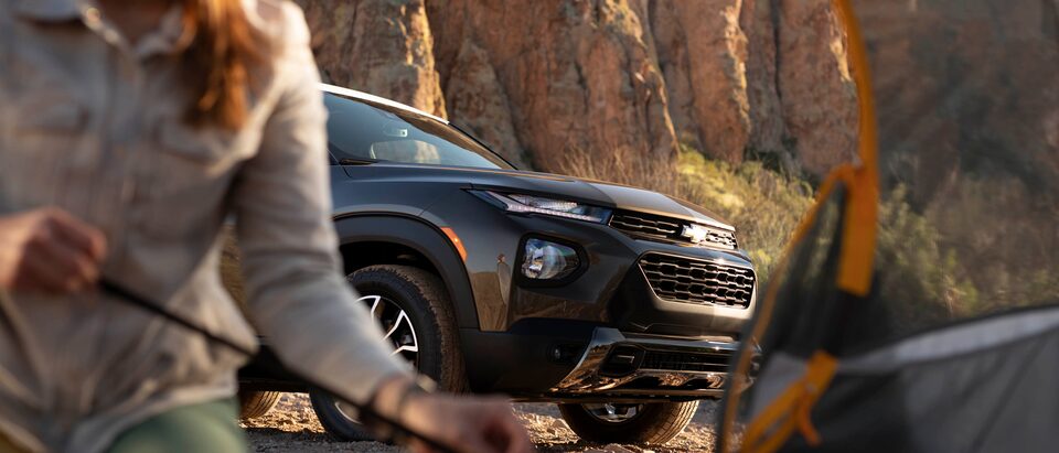 Seen from inside a tent through the open door, a woman is kneeling on the ground in front of the tent and a Zeus Bronze Metallic/Summit White Chevy Trailblazer ACTIV is behind her. In the background is a rocky cliff.