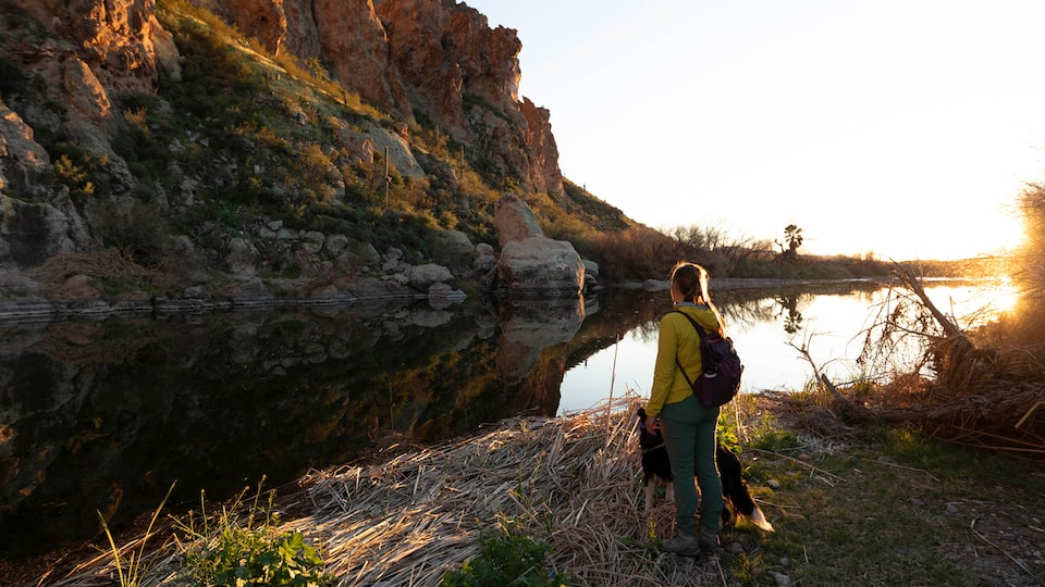 A woman wearing a backpack stands with her dog next to a pond in front of a rocky cliff.