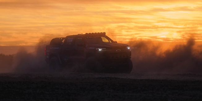 Chad Hall's racing Colorado ZR2 is seen in silhouette against a setting sun, with desert dust kicked up all around the vehicle.