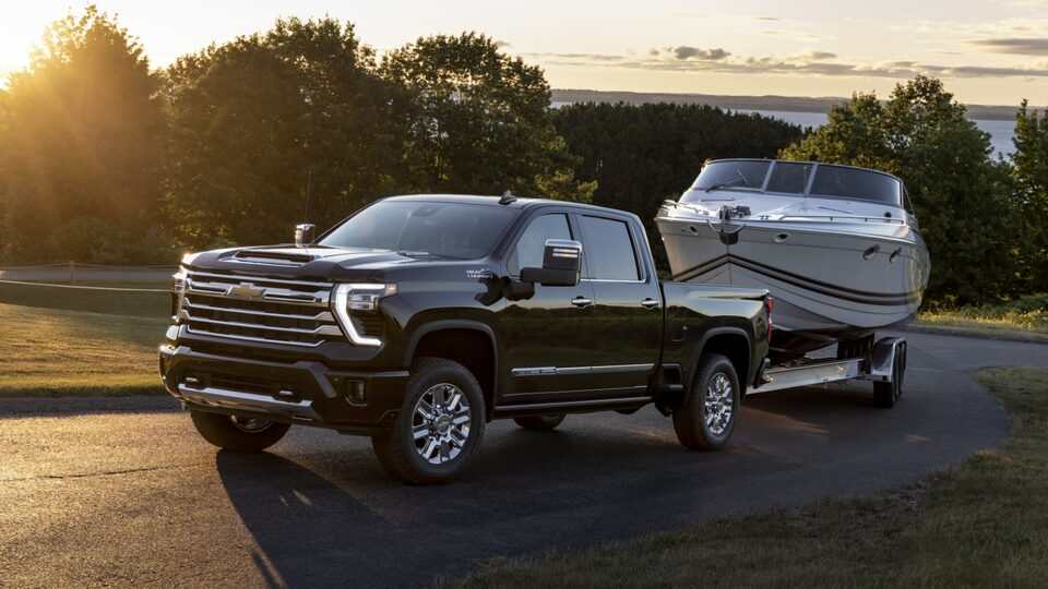 A black Silverado HD pulls a ski boat along a tree-lined road as the sun rises in the background.