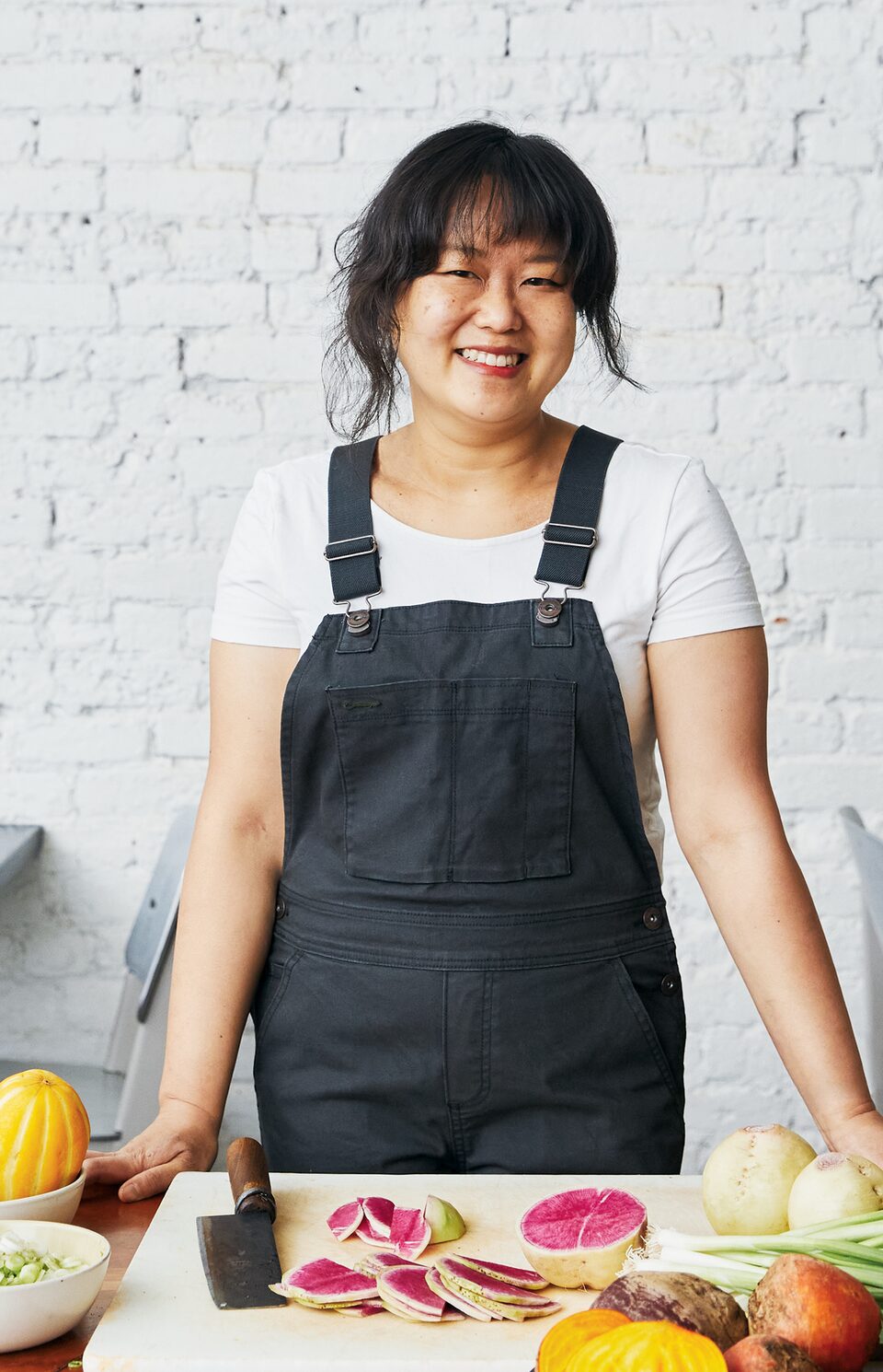 Chef Ji Hye Kim smiles as she stands at a long wooden table at her restaurant. On the table in front of her are a cutting board and cleaver along with a variety of fresh vegetables.