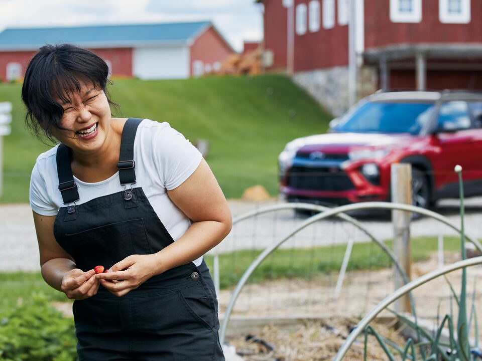 Chef Ji Hye Kim smiles as she stands in a garden with farm buildings and a red Chevy Trailblazer RS in the background.