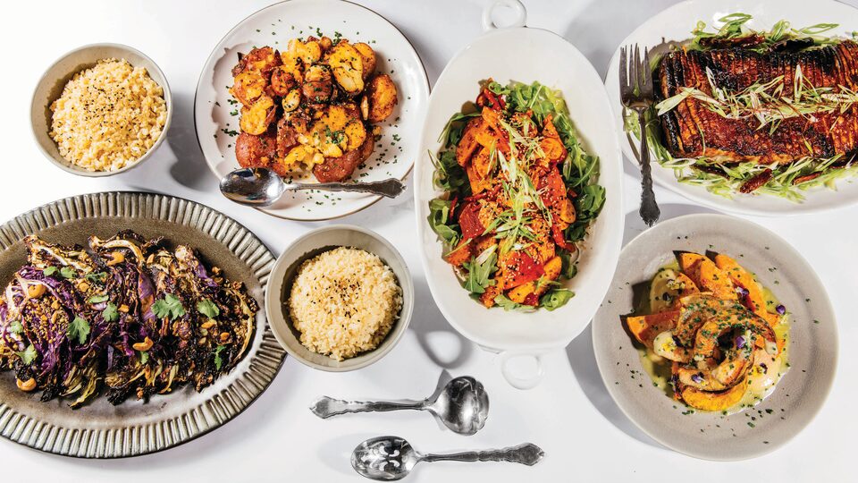 Looking down from above, several plates and platters filled with art fully arranged, colorful food sit on a white tabletop.