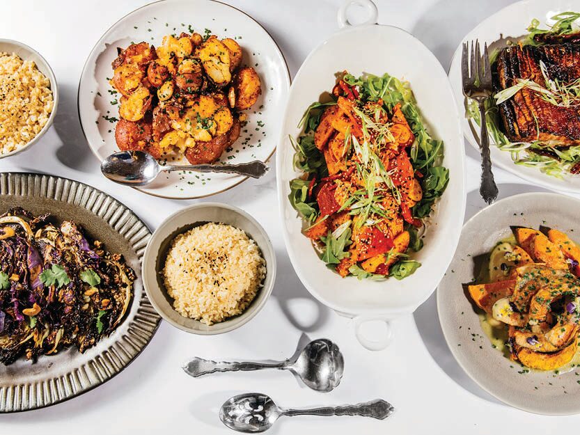 Looking down from above, several plates and platters filled with art fully arranged, colorful food sit on a white tabletop.