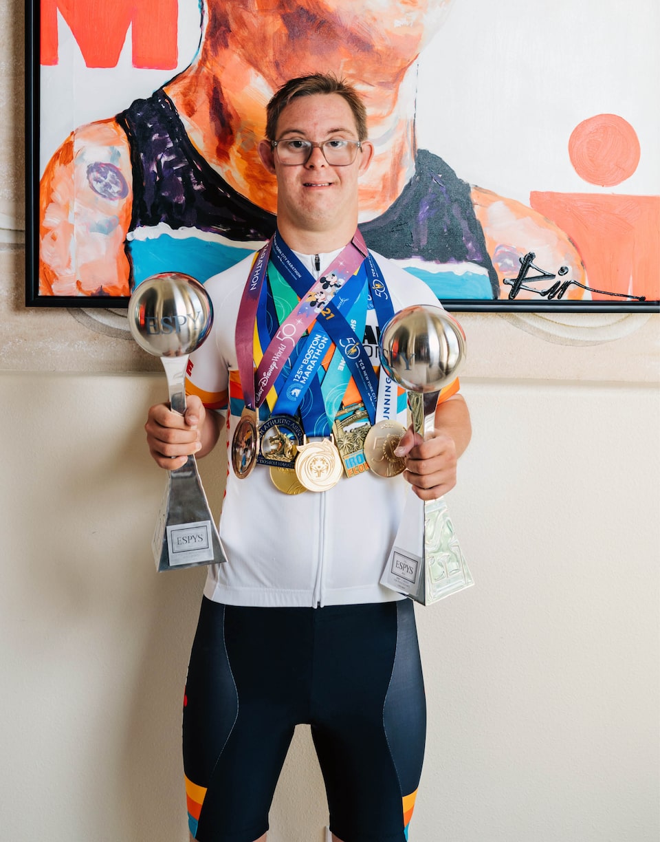 Chris Nikic, luciendo muchas medallas y sosteniendo trofeos, está de pie frente a un póster de sí mismo.