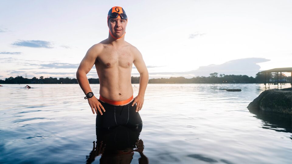 Chris Nikic se encuentra de pie, con las manos sobre las caderas, en las aguas superficiales de un lago. Usa un gorro de natación y su traje de neopreno bajado de sus brazos.