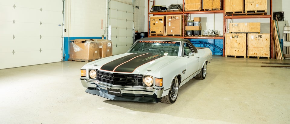 A 1972 El Camino, gray with black racing stripes on the hood, sits in a warehouse building.