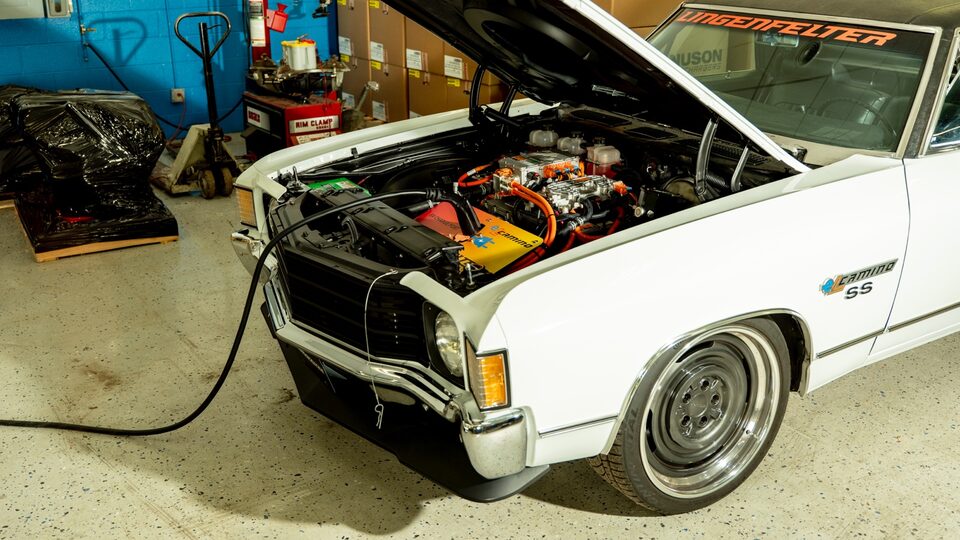 A 1972 El Camino is parked in a warehouse with its hood propped up. Below the hood are all the components of the electric engine.