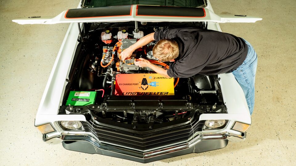 Seen from above, someone leans under the open hood of a 1972 El Camino and works on the battery-electric components inside.