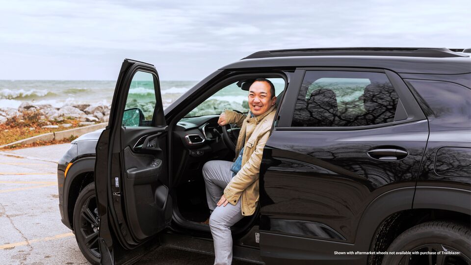 Erik Kennedy gira y sonríe a la cámara desde la ventanilla abierta del lado del conductor de su Trailblazer RS 2021 negra, que está estacionada en un lote con vista al lago Michigan.
